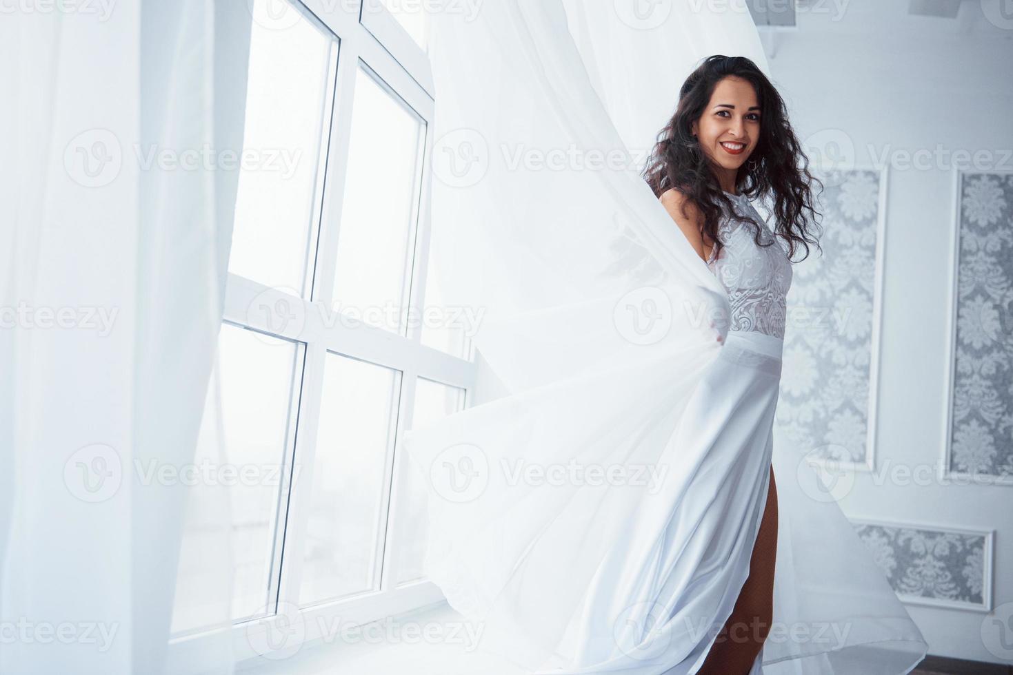 oprechte glimlach. mooie vrouw in witte jurk staat in witte kamer met daglicht door de ramen foto