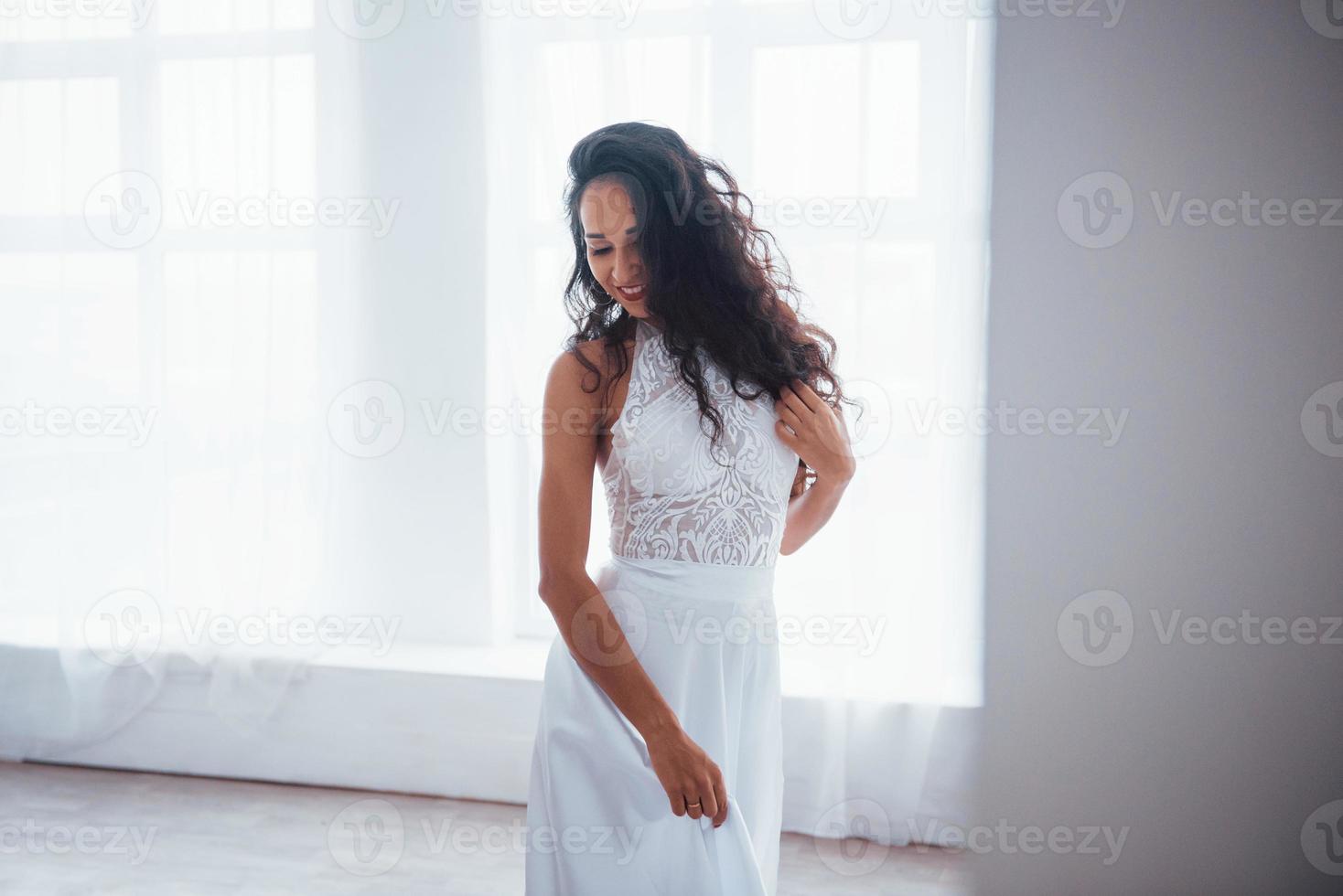 prachtig portret. mooie vrouw in witte jurk staat in witte kamer met daglicht door de ramen foto