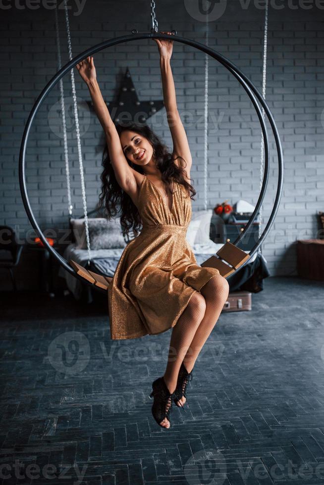 de bovenkant raken. mooie vrouw in goudkleurige jurk zit op de stijlvolle cirkelvormige bank aan de ketting. luxe kamer foto