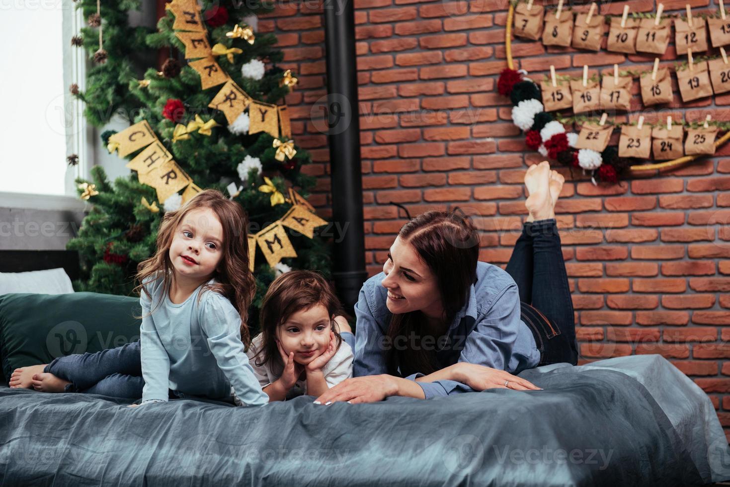 glimlachen en plezier hebben. moeder en twee kinderen die op het bed liggen met een kamer die versierd is met een kerstboom foto