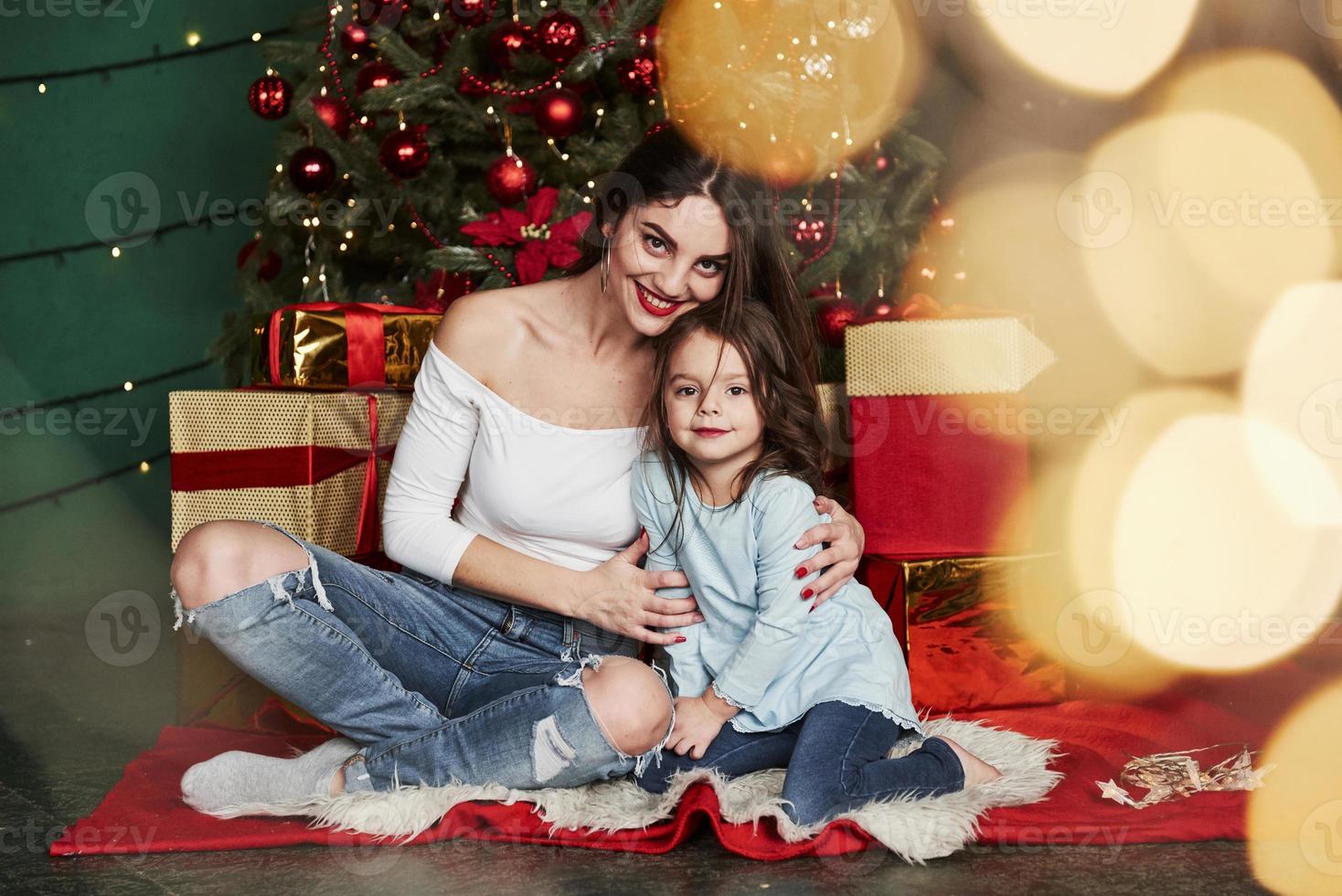 vakantie conceptie. vrolijke moeder en dochter zitten in de buurt van de kerstboom die erachter zit. schattig portret foto