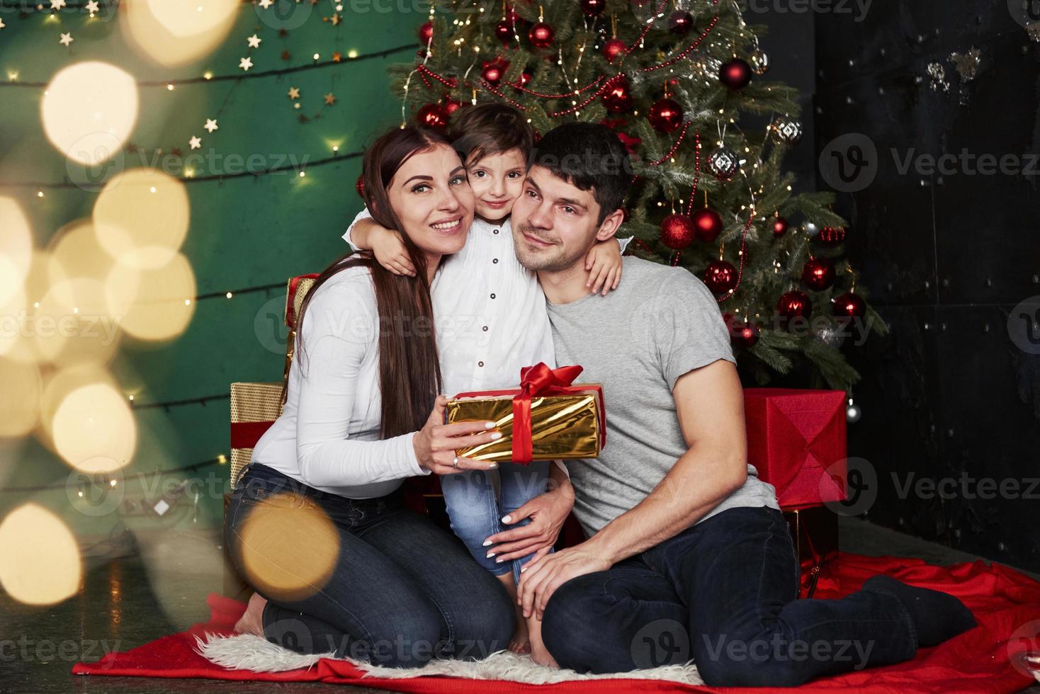 lieve knuffels. mooie familie zit in de buurt van de kerstboom met geschenkdozen op winteravond, genietend van de tijd die we samen doorbrengen foto