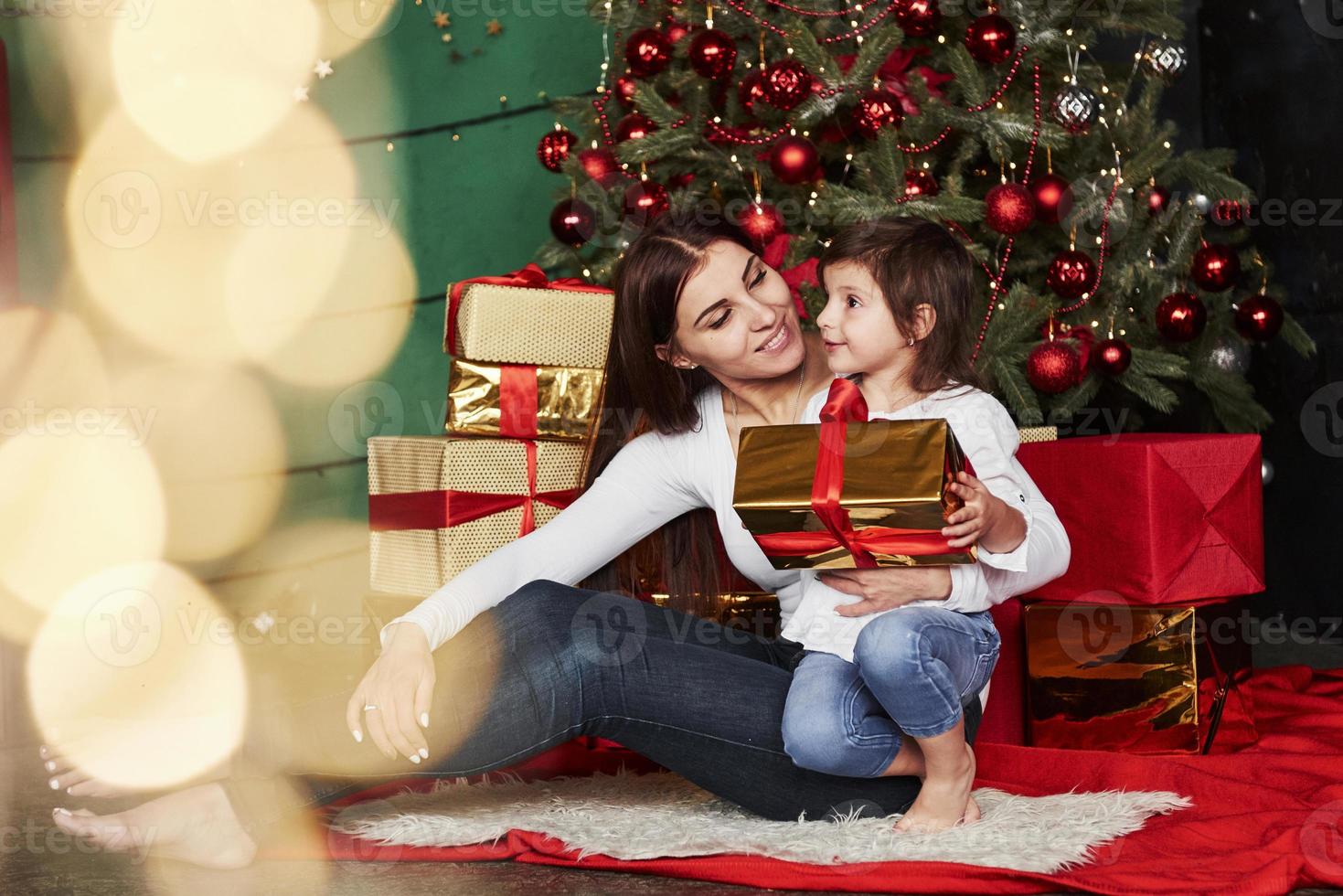 vakantie is mensen verenigen. vrolijke moeder en dochter zitten in de buurt van de kerstboom die erachter zit. schattig portret foto