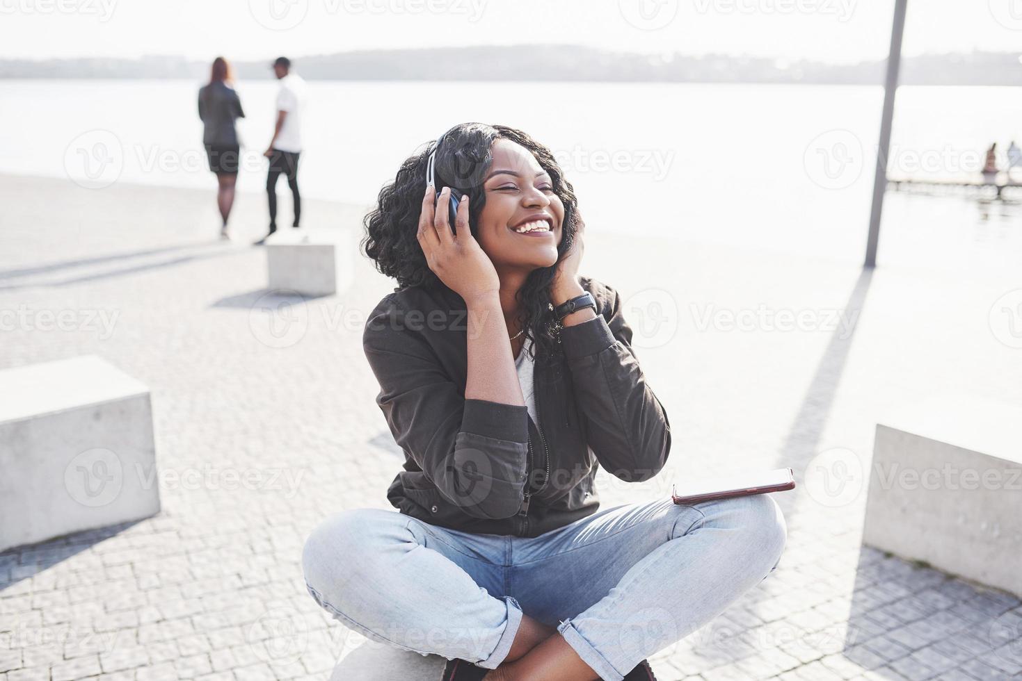 portret van een mooi jong mooi Afrikaans Amerikaans meisje zittend op het strand of het meer en luisterend naar muziek in haar koptelefoon foto