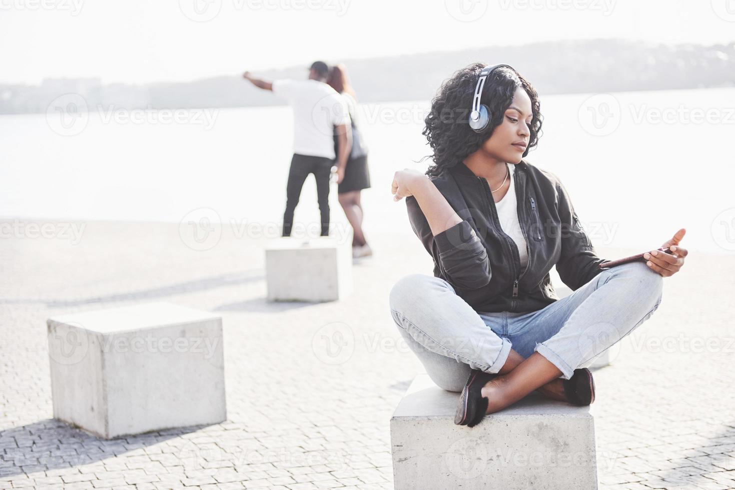 portret van een mooi jong mooi Afrikaans Amerikaans meisje zittend op het strand of het meer en luisterend naar muziek in haar koptelefoon foto