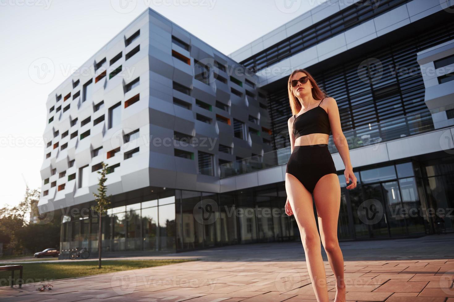 sexy jonge vrouw in zonnebril en zwart badpak staat tijdens zonsondergang op de achtergrond van de stadsgebouwen foto