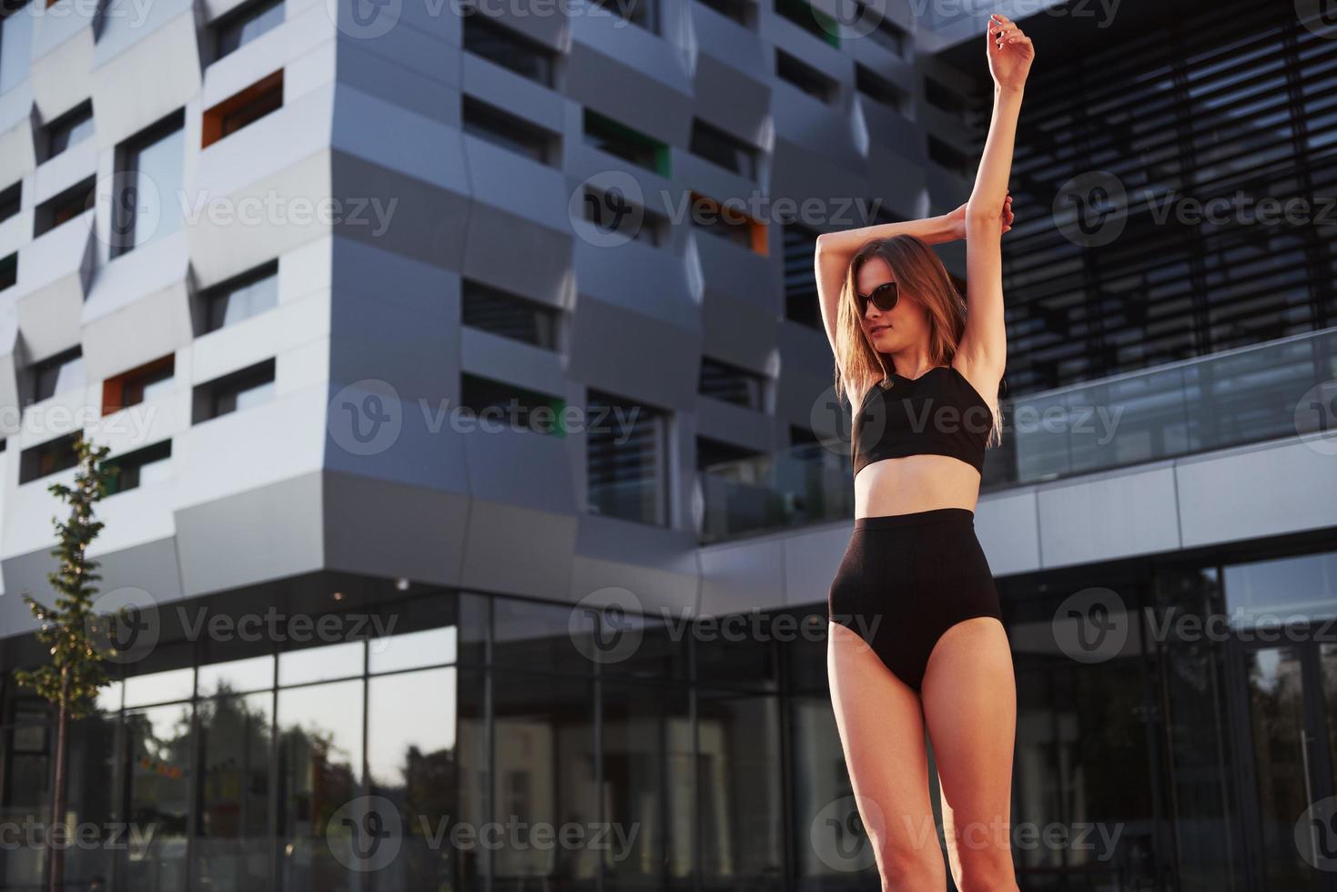 sexy jonge vrouw in zonnebril en zwart badpak staat tijdens zonsondergang op de achtergrond van de stadsgebouwen foto