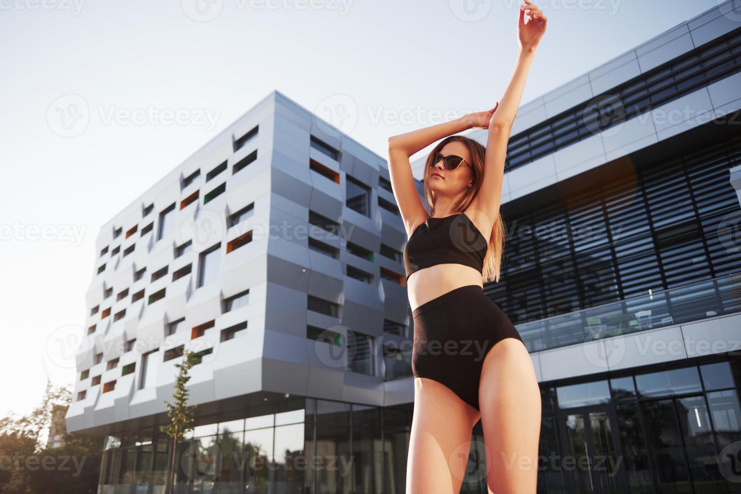 sexy jonge vrouw in zonnebril en zwart badpak staat tijdens zonsondergang op de achtergrond van de stadsgebouwen foto