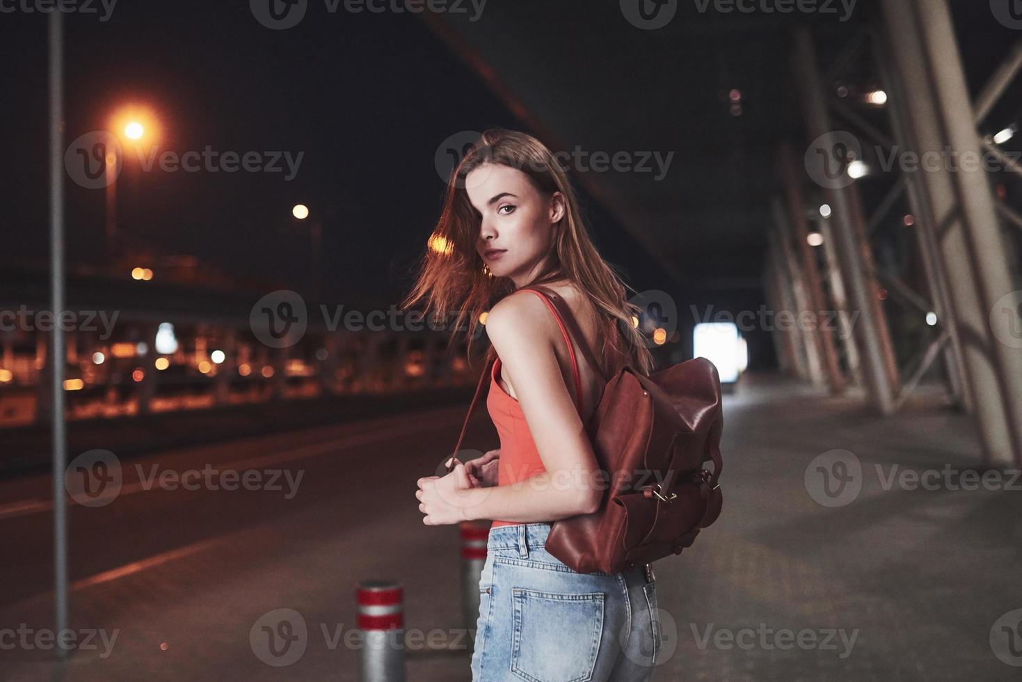 een jong mooi meisje met een rugzak achter haar schouders staat op een warme zomeravond op straat in de buurt van een luchthaven of een treinstation. ze is net aangekomen en wacht op een taxi of haar vrienden foto