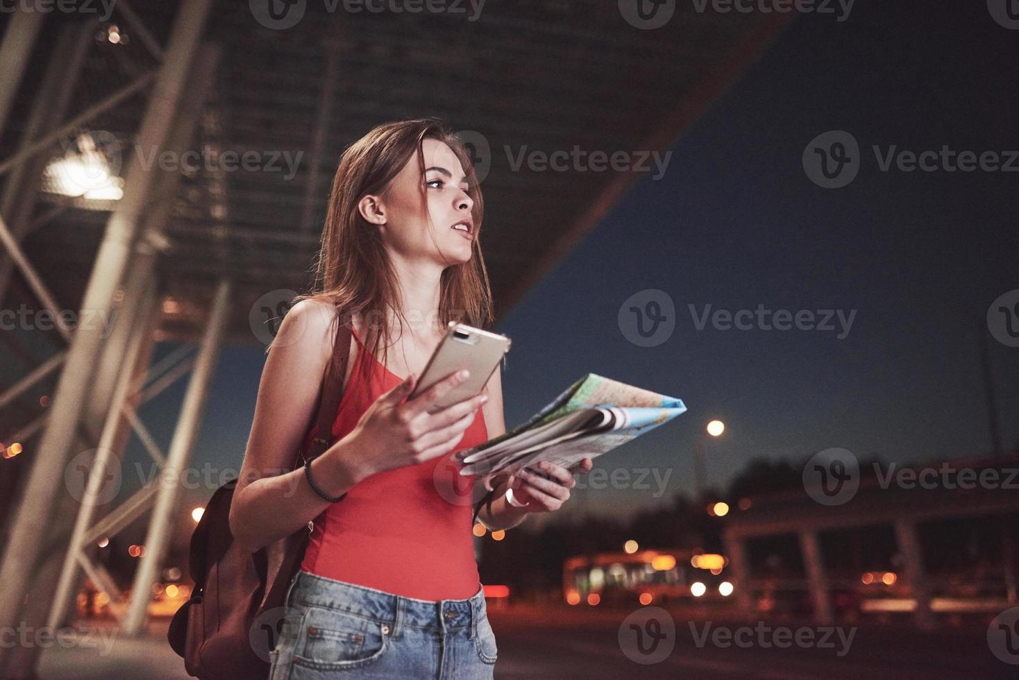 jong meisje kost 's nachts in de buurt van de terminal van de luchthaven of het station en het lezen van de stadsplattegrond en op zoek naar een hotel. schattige toerist met rugzakken bepalen het concept van reizen foto