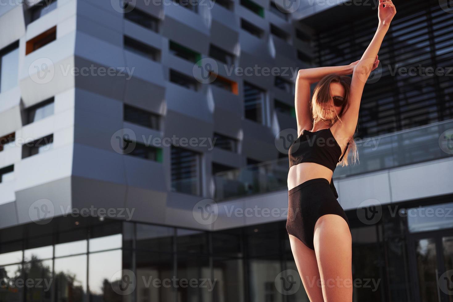 sexy jonge vrouw in zonnebril en zwart badpak staat tijdens zonsondergang op de achtergrond van de stadsgebouwen foto