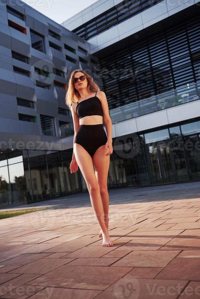 sexy jonge vrouw in zonnebril en zwart badpak staat tijdens zonsondergang op de achtergrond van de stadsgebouwen foto