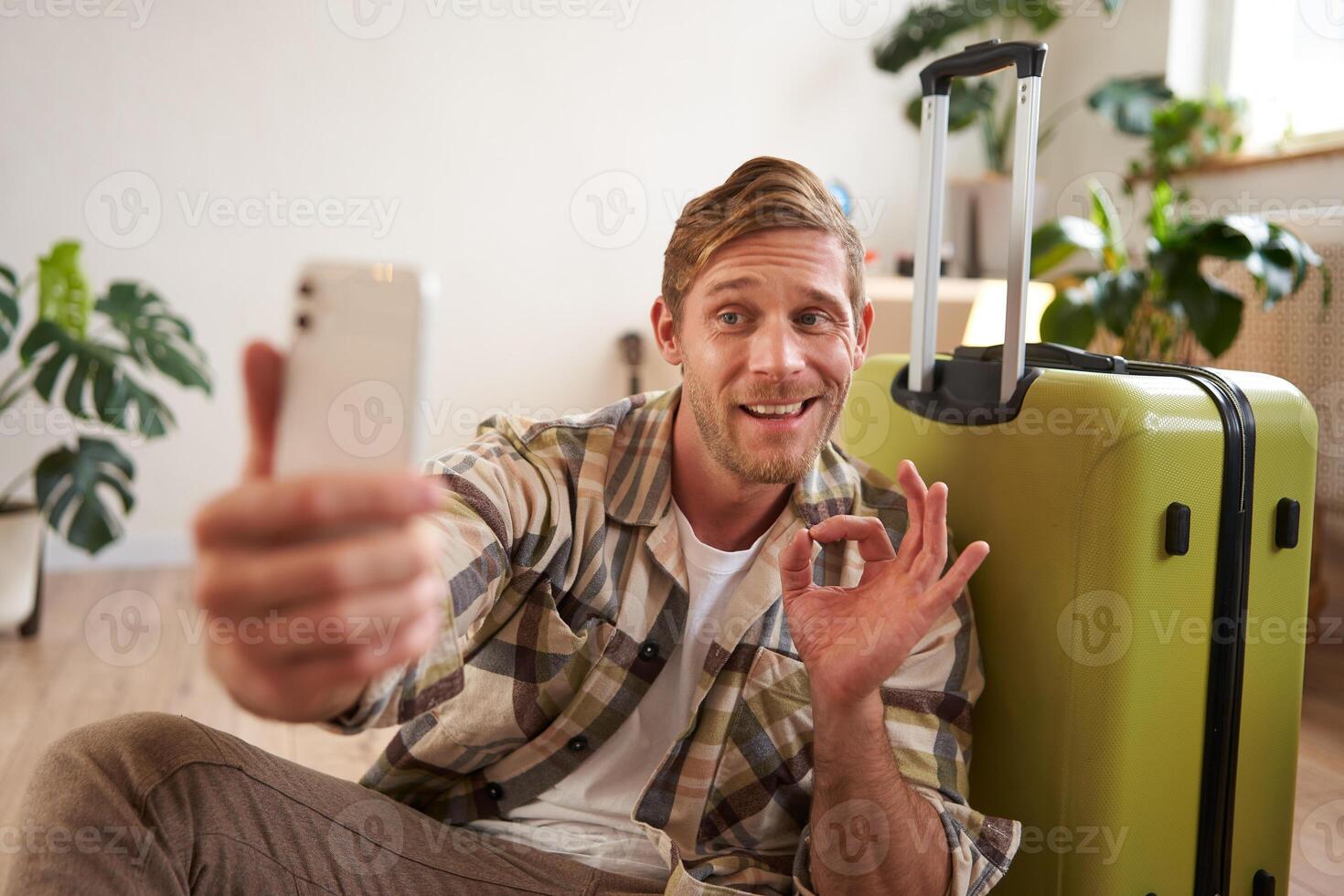 portret van knap glimlachen vent met koffer, duurt selfie Aan smartphone, shows OK teken, goedkeuren vakantie bestemming, berichten foto's Aan sociaal media account foto