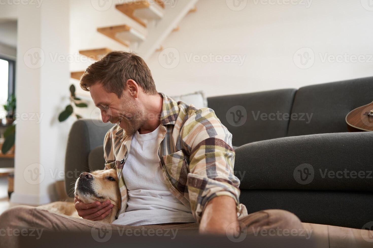 portret van gelukkig jong Mens, knuffelen en spelen met zijn hond, zittend Aan verdieping met bank, op zoek Bij gouden retriever foto