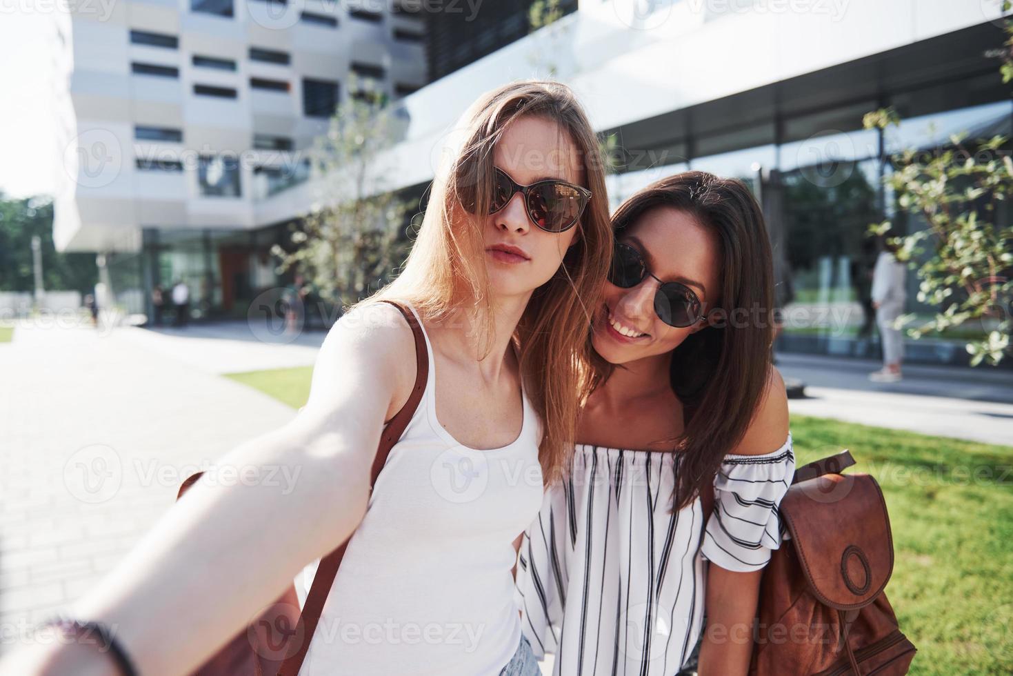 twee mooie gelukkige jonge student meisje met rugzak in de buurt van de campus van de universiteit en selfie foto maken. onderwijs en vrije tijd concept