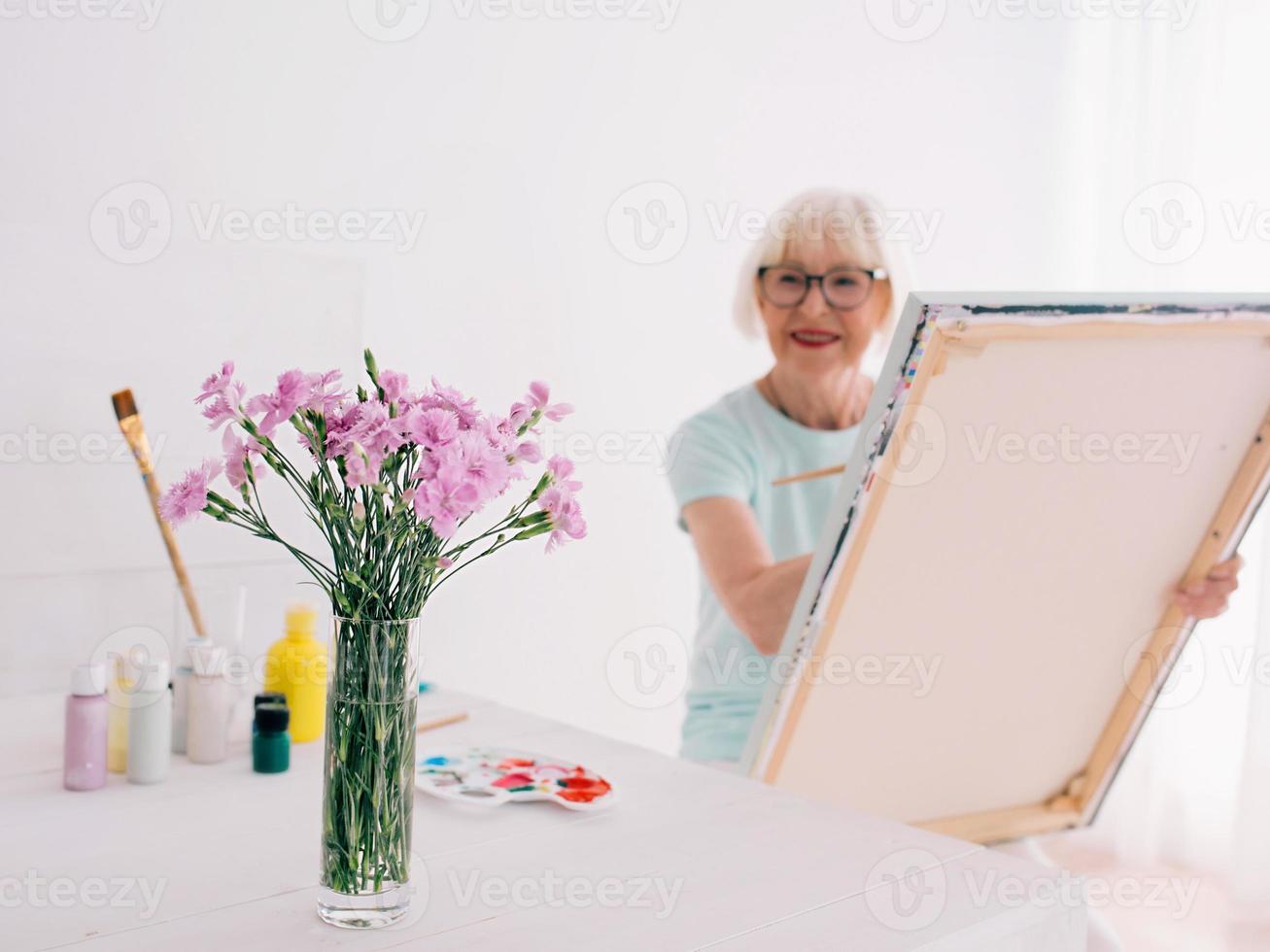 senior vrolijke vrouw kunstenaar in glazen met grijze haren schilderen bloemen in vaas. creativiteit, kunst, hobby, bezettingsconcept foto