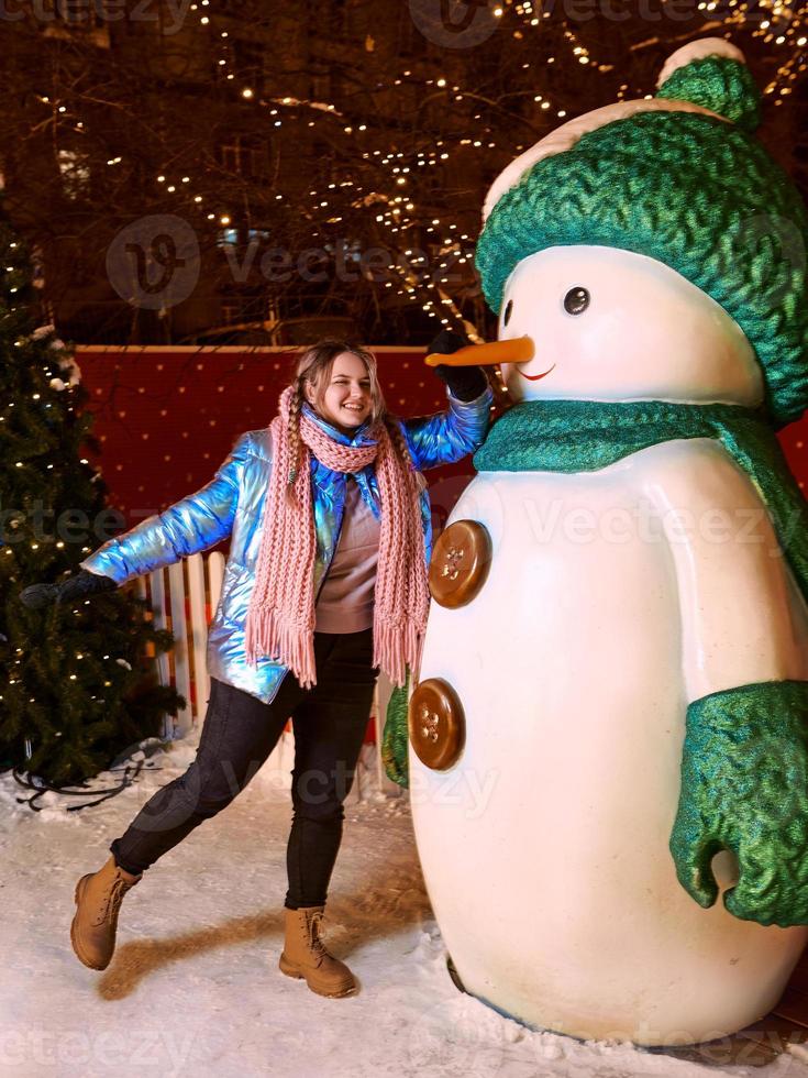 gelukkig lachende jonge blanke vrouw in sjaal, muts, jas, wanten door de sneeuwpop buiten. nieuwjaar, plezier, winterconcept foto