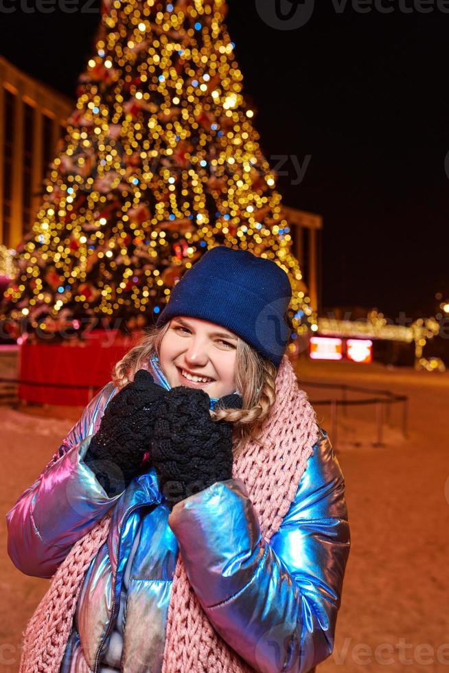 gelukkig lachende jonge blanke vrouw in sjaal, muts, jas, wanten bij de kerstboom buiten. nieuwjaar, plezier, winterconcept foto