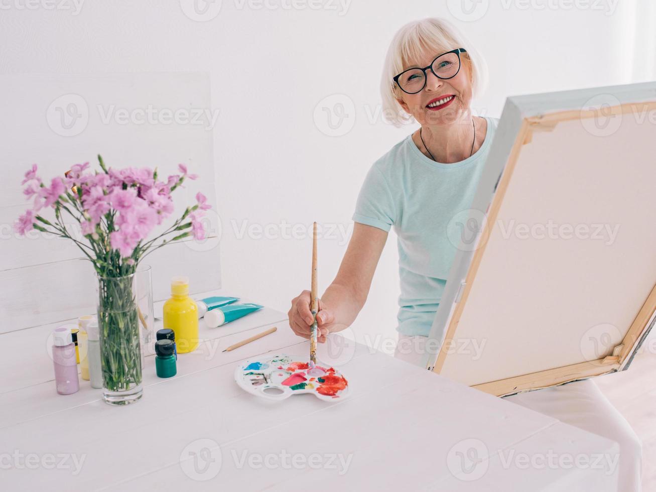 senior vrolijke vrouw kunstenaar in glazen met grijze haren schilderen bloemen in vaas. creativiteit, kunst, hobby, bezettingsconcept foto