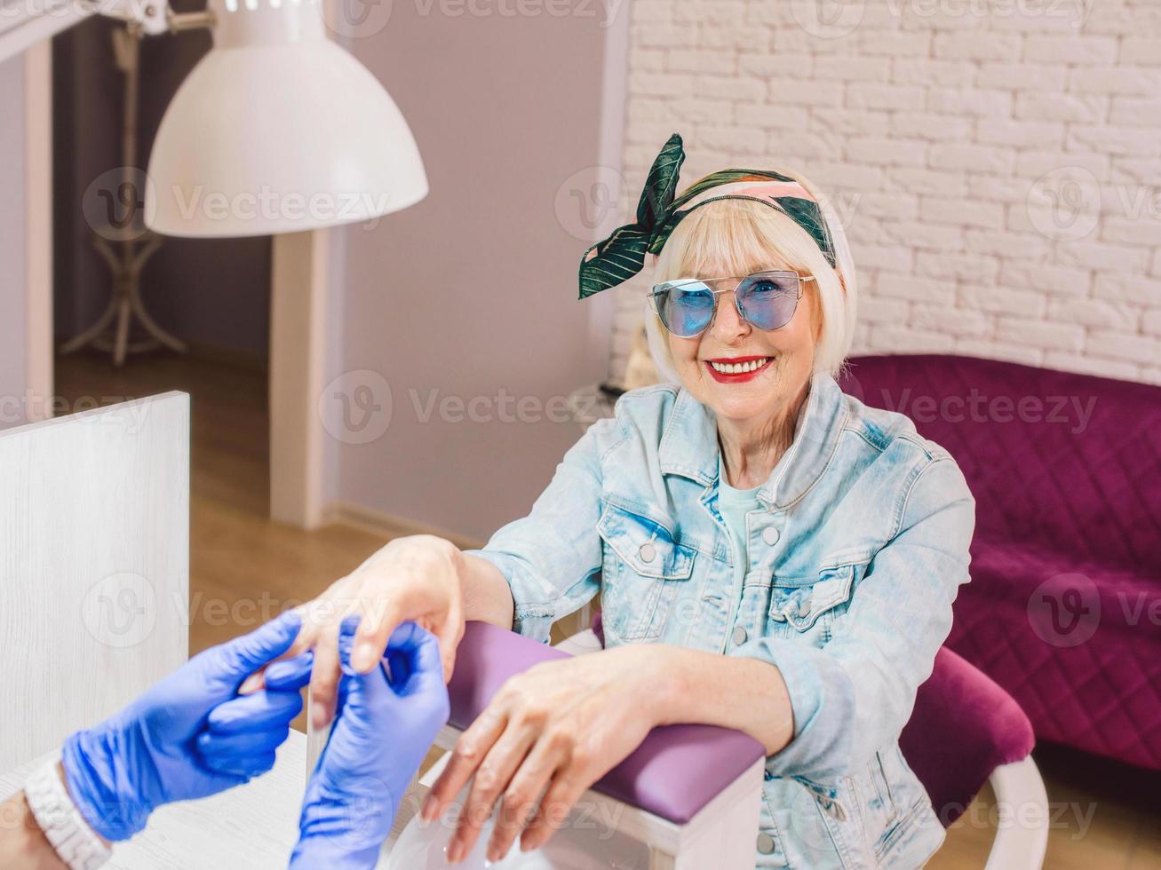 manicure meester in blauwe handschoenen afroming handen van oudere stijlvolle vrouw in blauwe zonnebril en spijkerjasje zittend op manicure salon foto