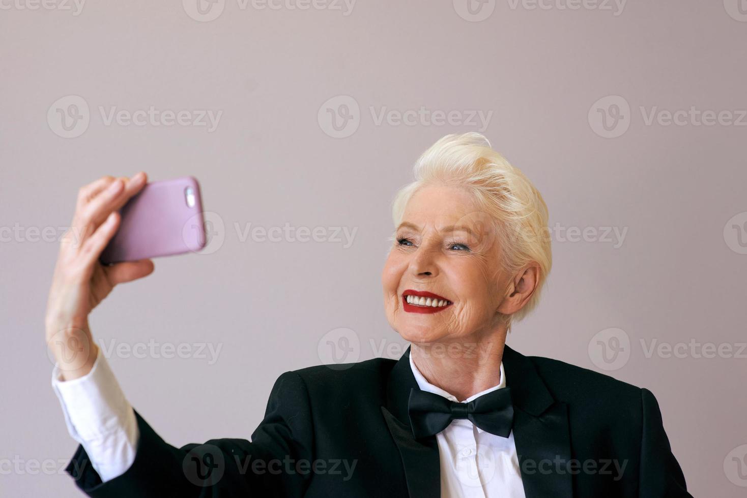 stijlvolle volwassen senior vrouw in smoking met mobiel videobellen of selfie maken. plezier, feest, stijl, levensstijl, bedrijf, technologie, feestconcept foto