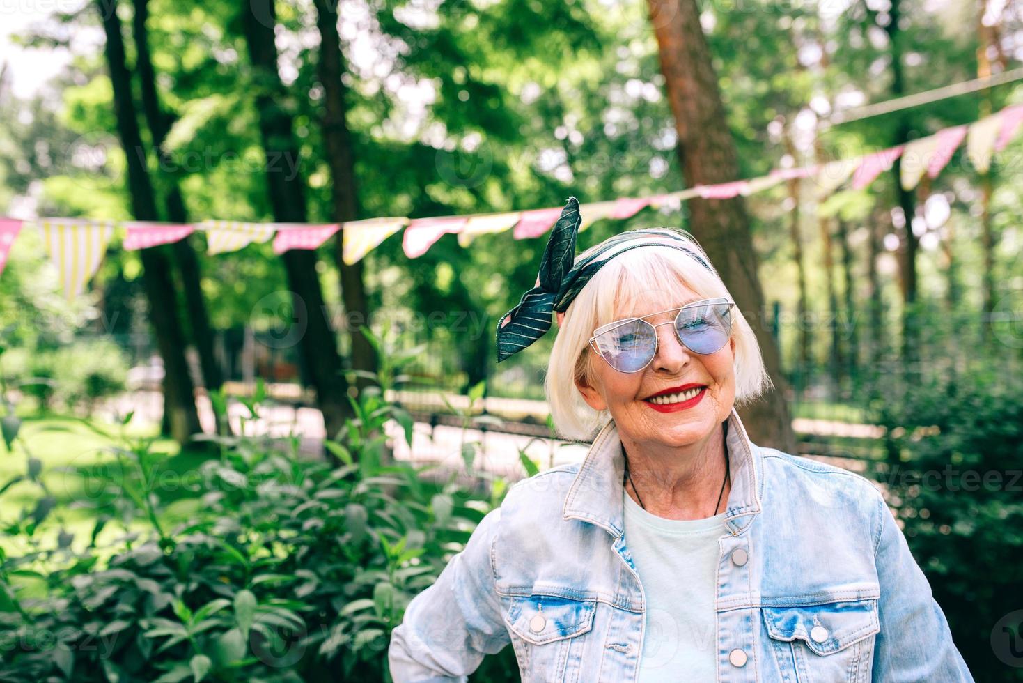 vrolijke senior stijlvolle vrouw met grijs haar en in blauwe bril en denim vakantiemeester buitenshuis. vakantie, feest, anti-leeftijd, leuk concept foto