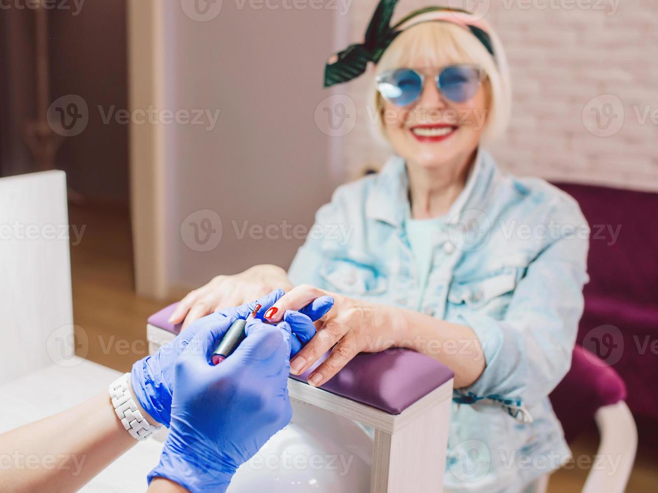 manicure meester in blauwe handschoenen afroming handen van oudere stijlvolle vrouw in blauwe zonnebril en spijkerjasje zittend op manicure salon foto