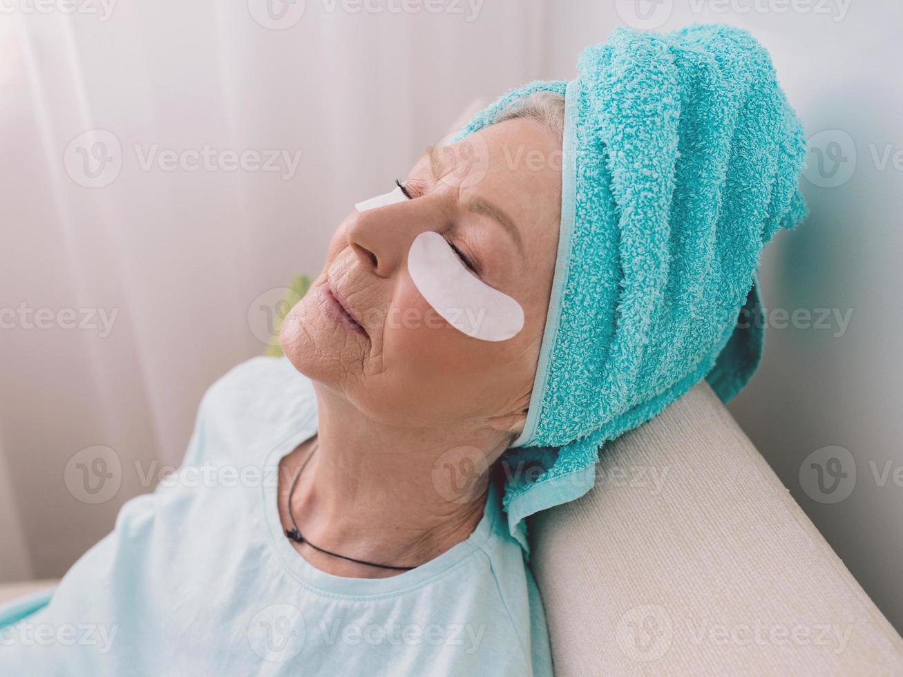 senior blanke stijlvolle vrouw die op een coach ligt met een blauwe handdoek op haar had en collageen ooglapjes. anti leeftijd, gezonde levensstijl, foto