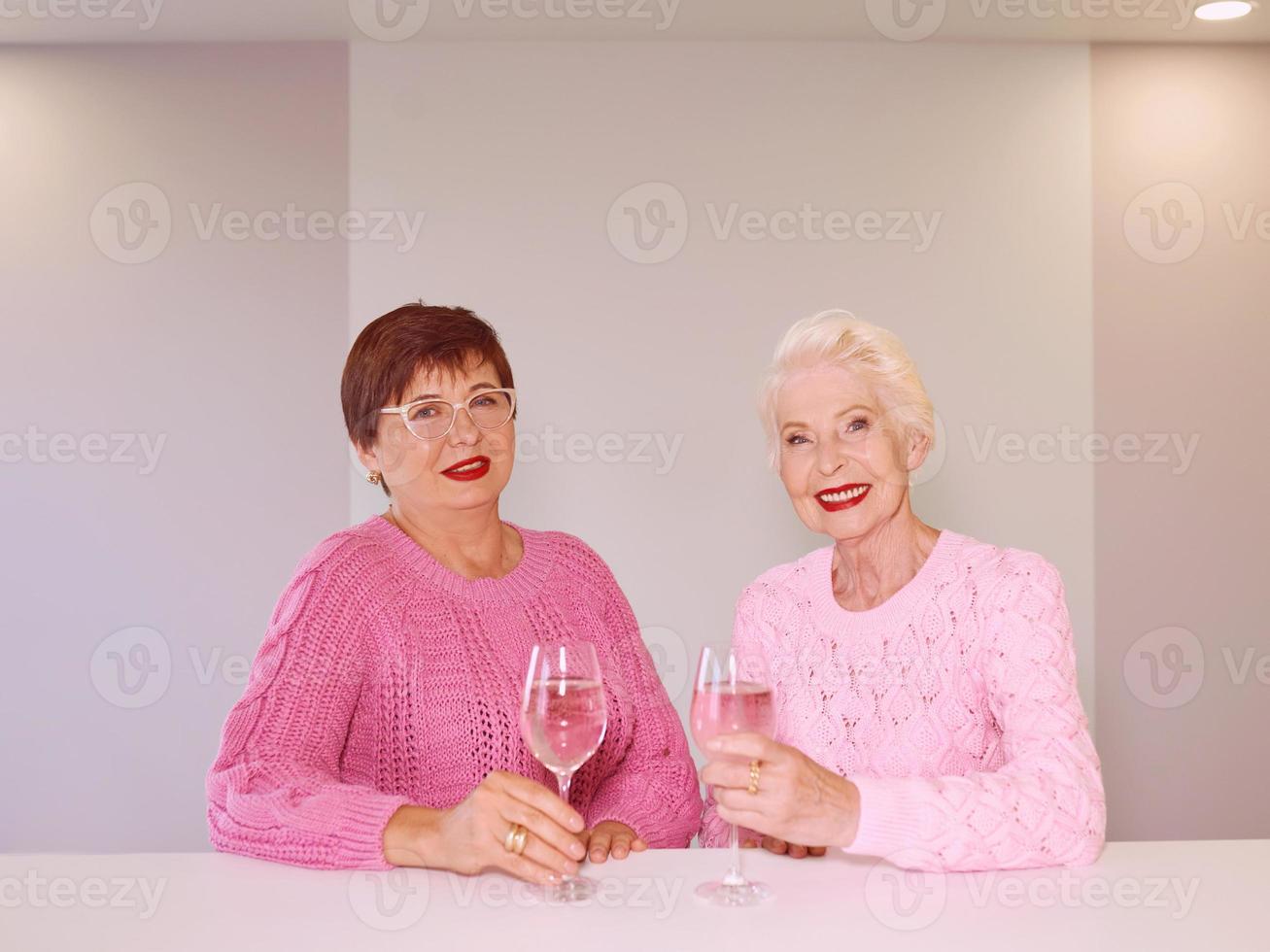 twee stijlvolle senior vrouwen in roze truien drinken rose wijn bij moderne keuken roddelen. vriendschap, praten, roddels, evenement, relaties, nieuws, familieconcept foto