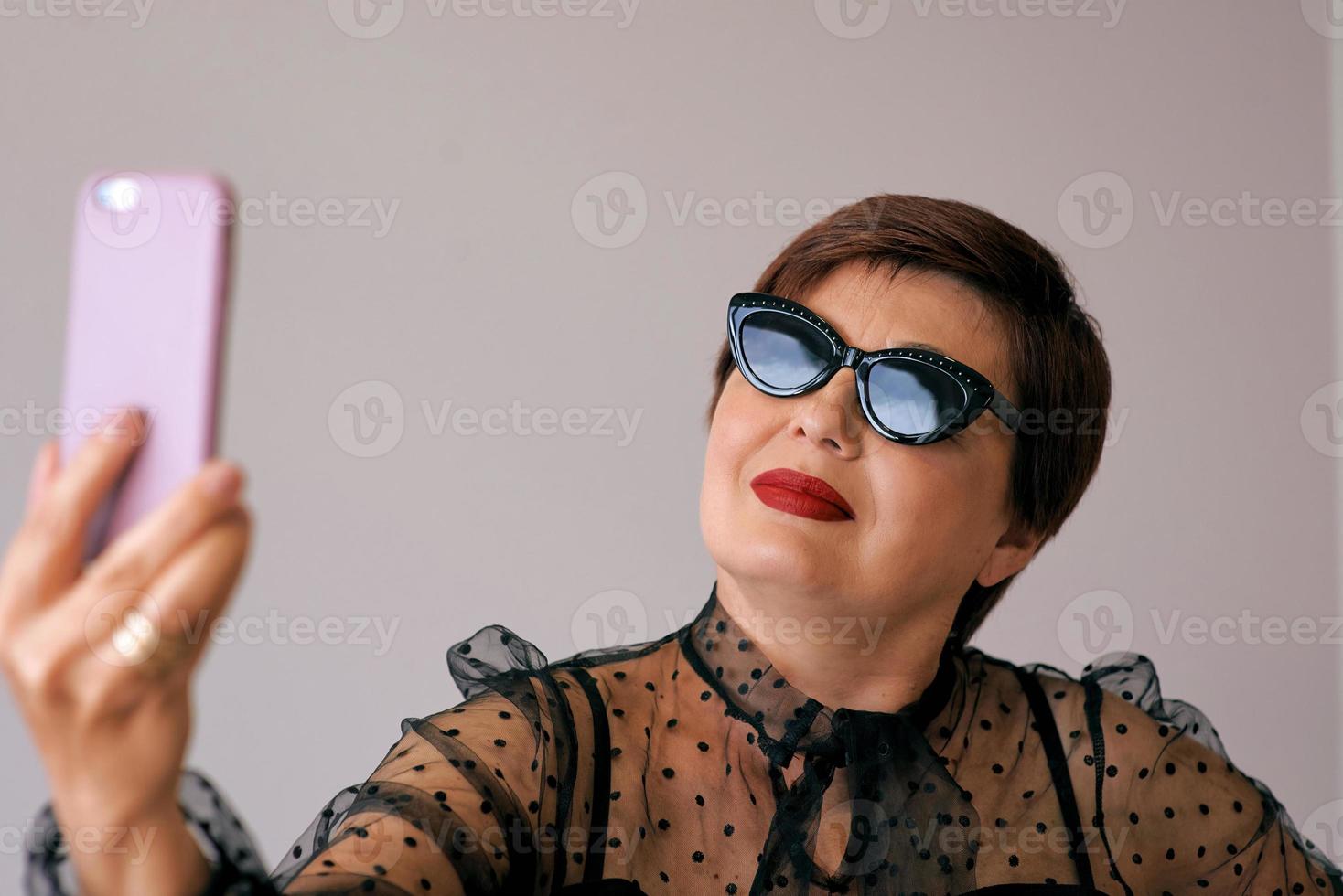 stijlvolle volwassen senior vrouw in rode blouse met mobiel videobellen of selfie maken. plezier, feest, stijl, levensstijl, technologie, feestconcept foto