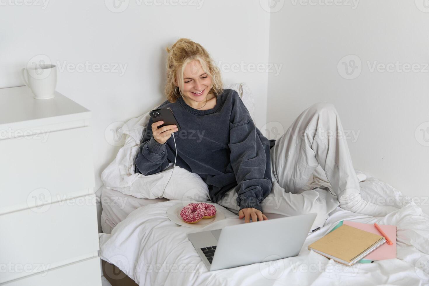 portret van glimlachen openhartig vrouw, aan het liegen in bed met donut, gebruik makend van smartphone en laptop, resting Bij huis in slaapkamer, aan het kijken TV tonen of chatten online foto