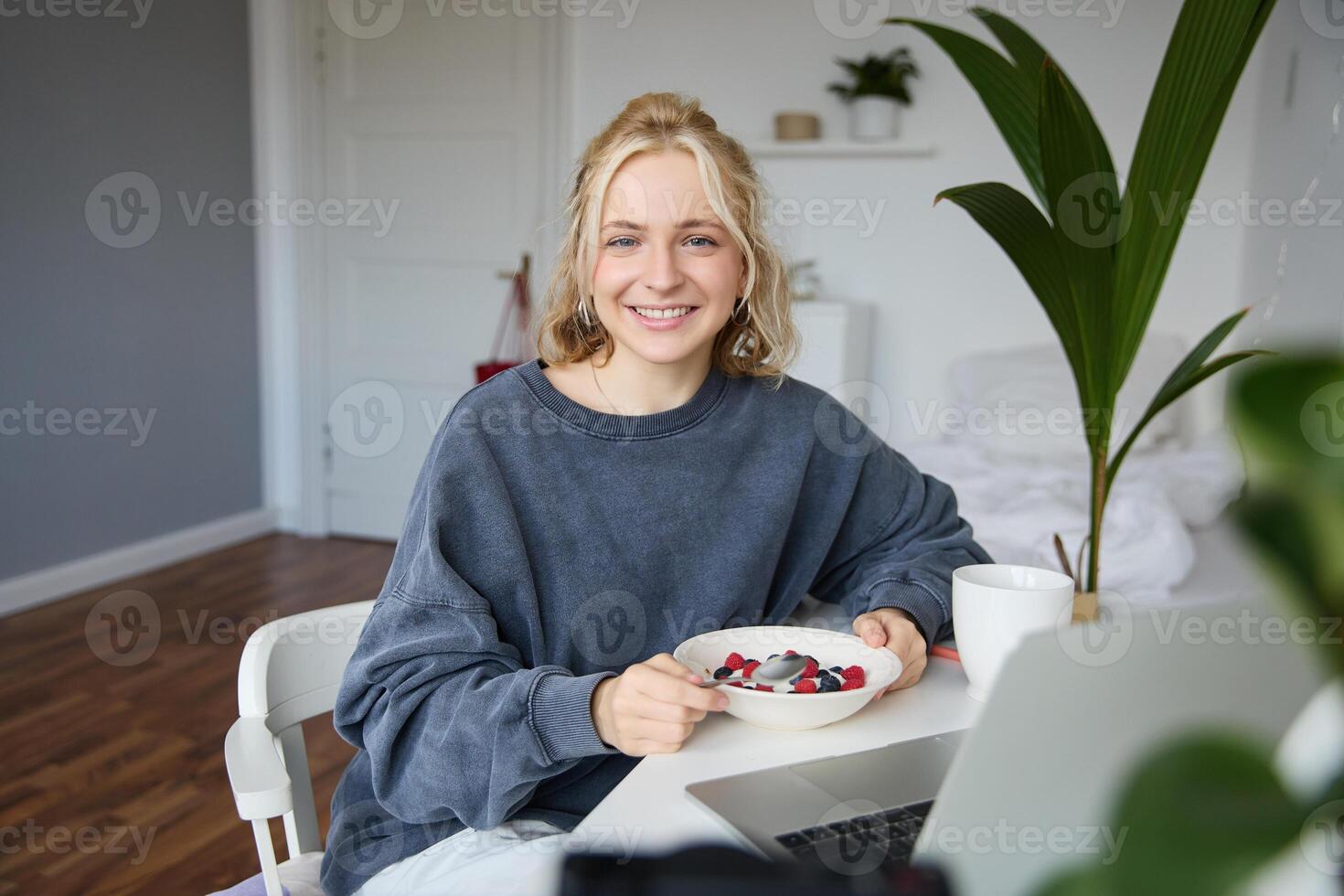 portret van jong vrouw aan het eten gezond maaltijd in een kamer, aan het kijken s Aan laptop, hebben lunch in voorkant van computer, glimlachen en op zoek gelukkig Bij camera foto