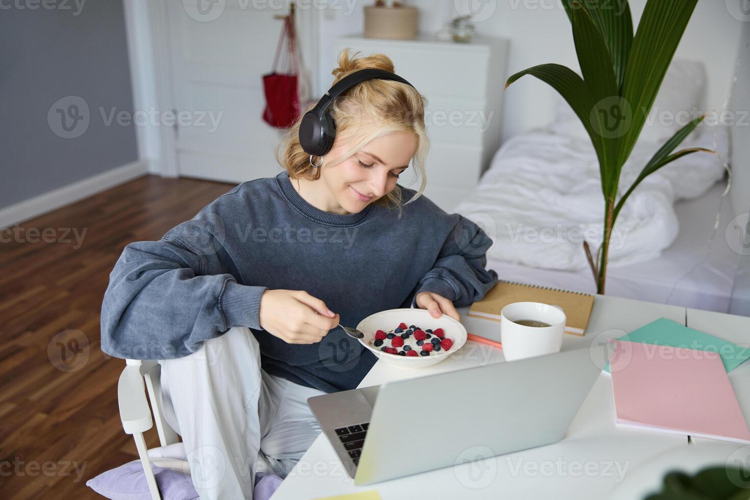 portret van glimlachen jong vrouw, aan het kijken TV tonen in koptelefoon, aan het eten ontbijt en op zoek Bij laptop scherm foto