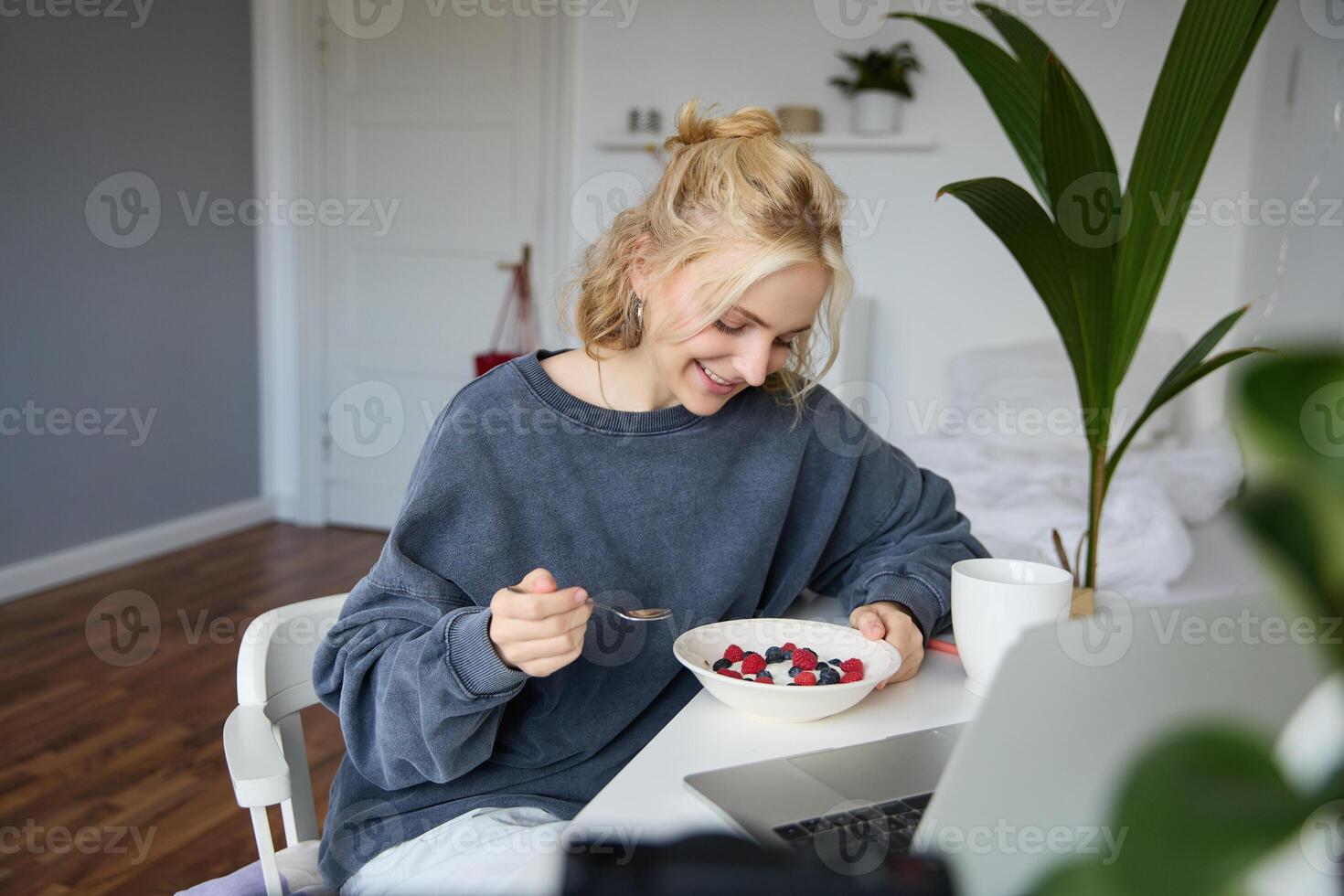 portret van glimlachen blond jong vrouw, aan het eten in voorkant van laptop, aan het kijken s online terwijl hebben ontbijt, genieten van nagerecht, zittend in slaapkamer foto