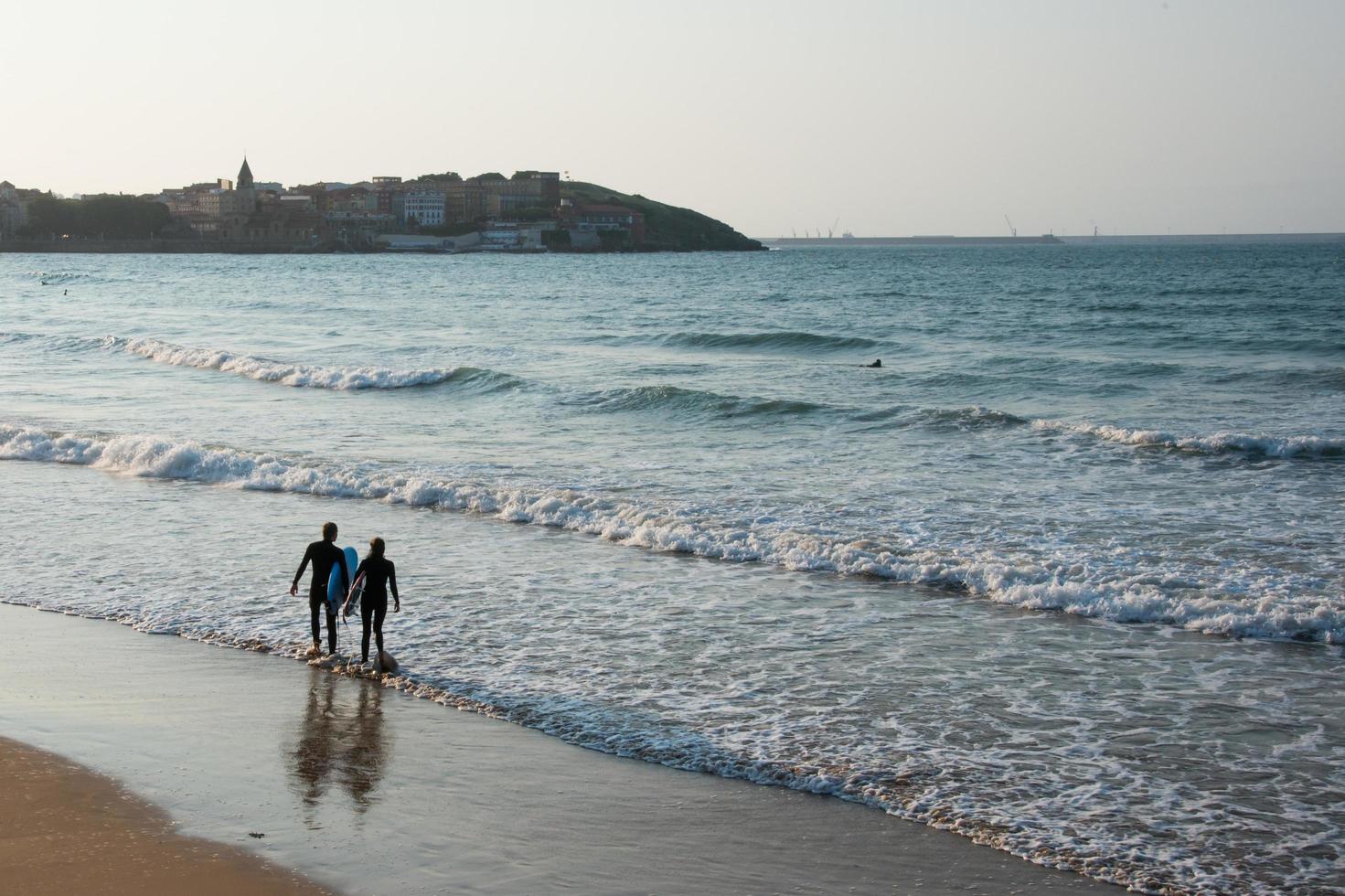 jong stel gezien vanaf hun rug het water in te gaan om te surfen op het strand van Gijon bij zonsondergang. uitzicht op de stad in de verte. asturië, spanje. foto