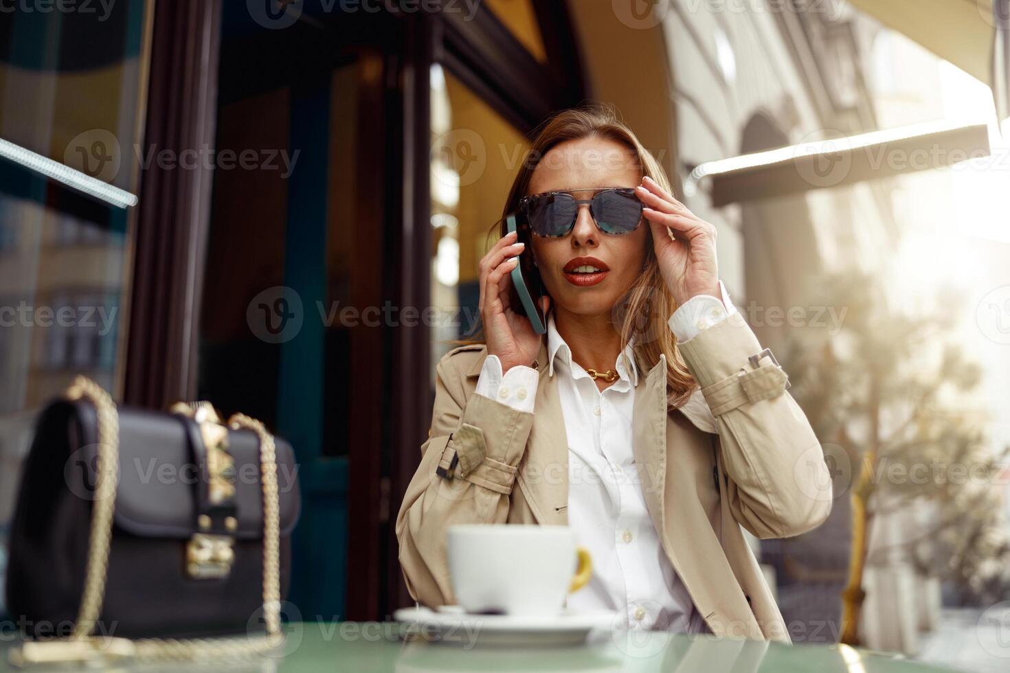 mooi vrouw vervelend zonnebril zittend Bij cafe terras pratend Aan telefoon terwijl drinken koffie foto