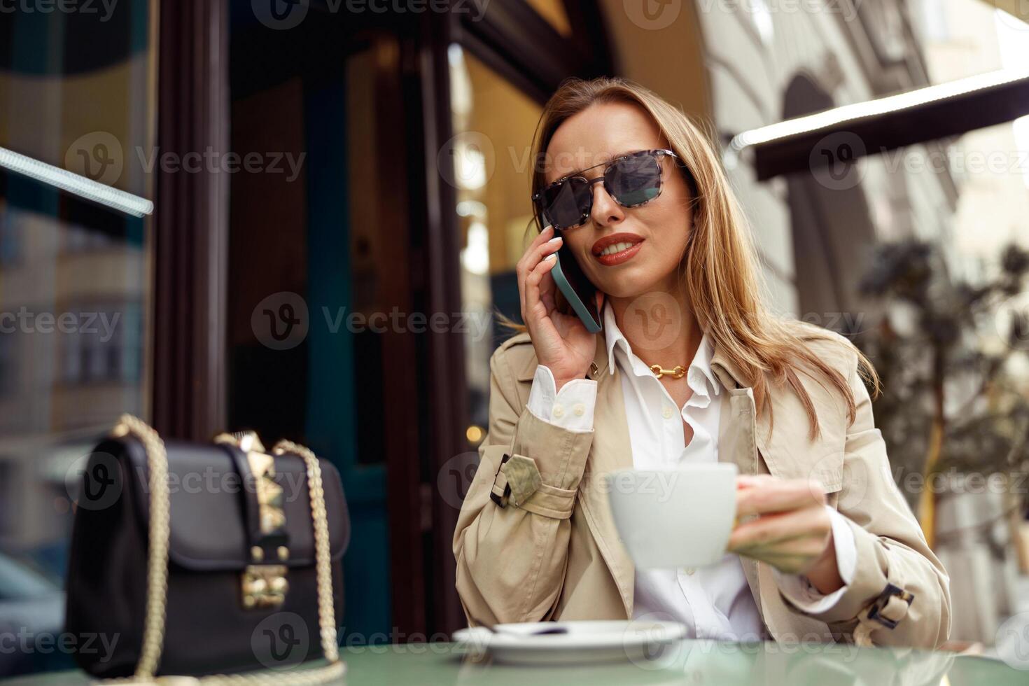 mooi vrouw vervelend zonnebril zittend Bij cafe terras pratend Aan telefoon terwijl drinken koffie foto