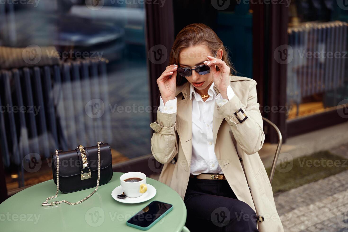 elegant jong vrouw vervelend zonnebril terwijl drinken koffie Aan restaurant terras foto