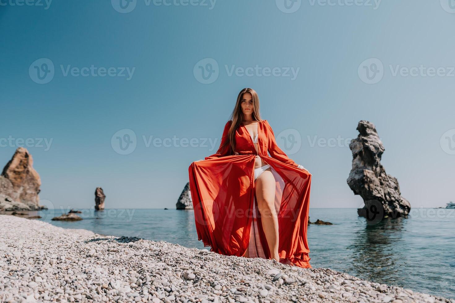 vrouw reizen zee. gelukkig toerist in rood jurk genieten nemen afbeelding buitenshuis voor herinneringen. vrouw reiziger poseren Aan de rots Bij zee baai omringd door vulkanisch bergen, sharing reizen avontuur reis foto