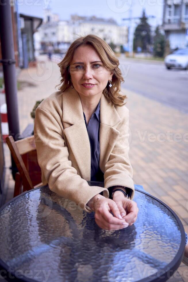 jong volwassen vrouw in jeans en zand jas Bij een tafel in een cafe Aan een voorjaar dag foto