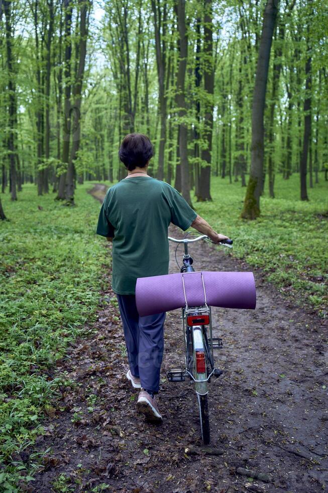 ouderen vrouw wandelen in voorjaar Woud met fiets en yoga mat foto