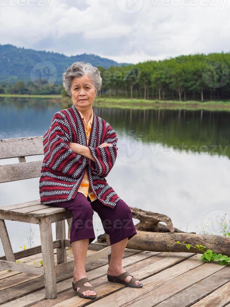 portret van ouderen Aziatisch vrouw armen gekruiste en op zoek Bij de camera terwijl zittend Aan een houten stoel kant van de meer met bergen achtergrond. concept van oud mensen en ontspanning foto
