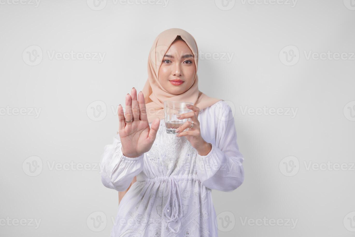 echt jong Aziatisch moslim vrouw vervelend wit jurk en hijab tonen haar palm naar maken een hou op gebaar terwijl Holding een kop van water, staand over- geïsoleerd wit achtergrond. Ramadan concept foto