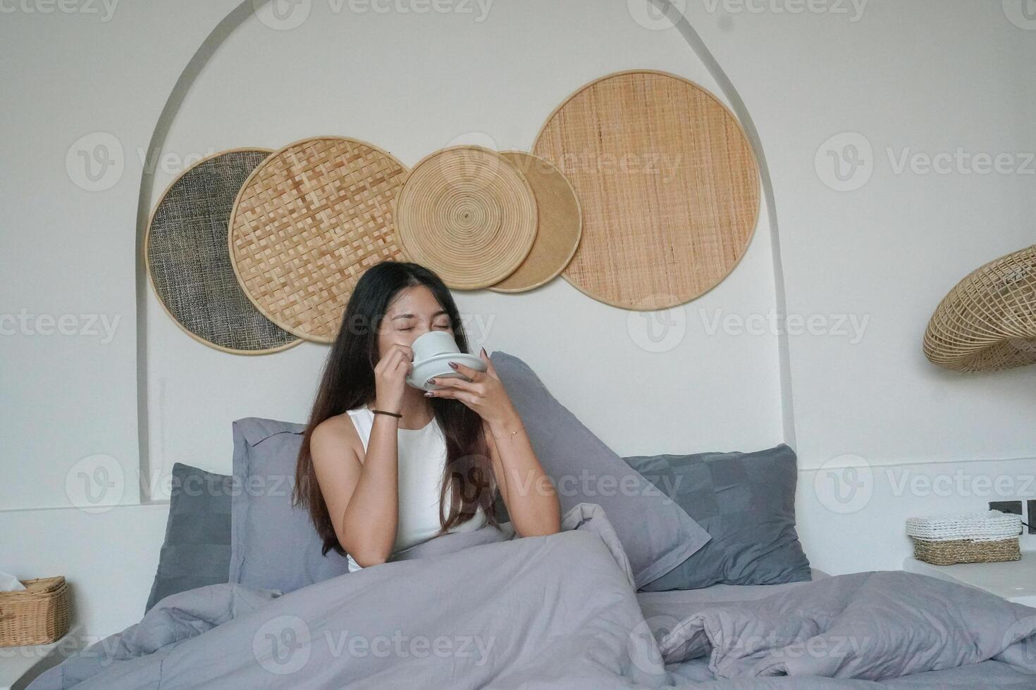 jong Aziatisch vrouw drinken ochtend- koffie of thee in een kop zittend Aan bed na ontwaken omhoog vervelend wit top foto