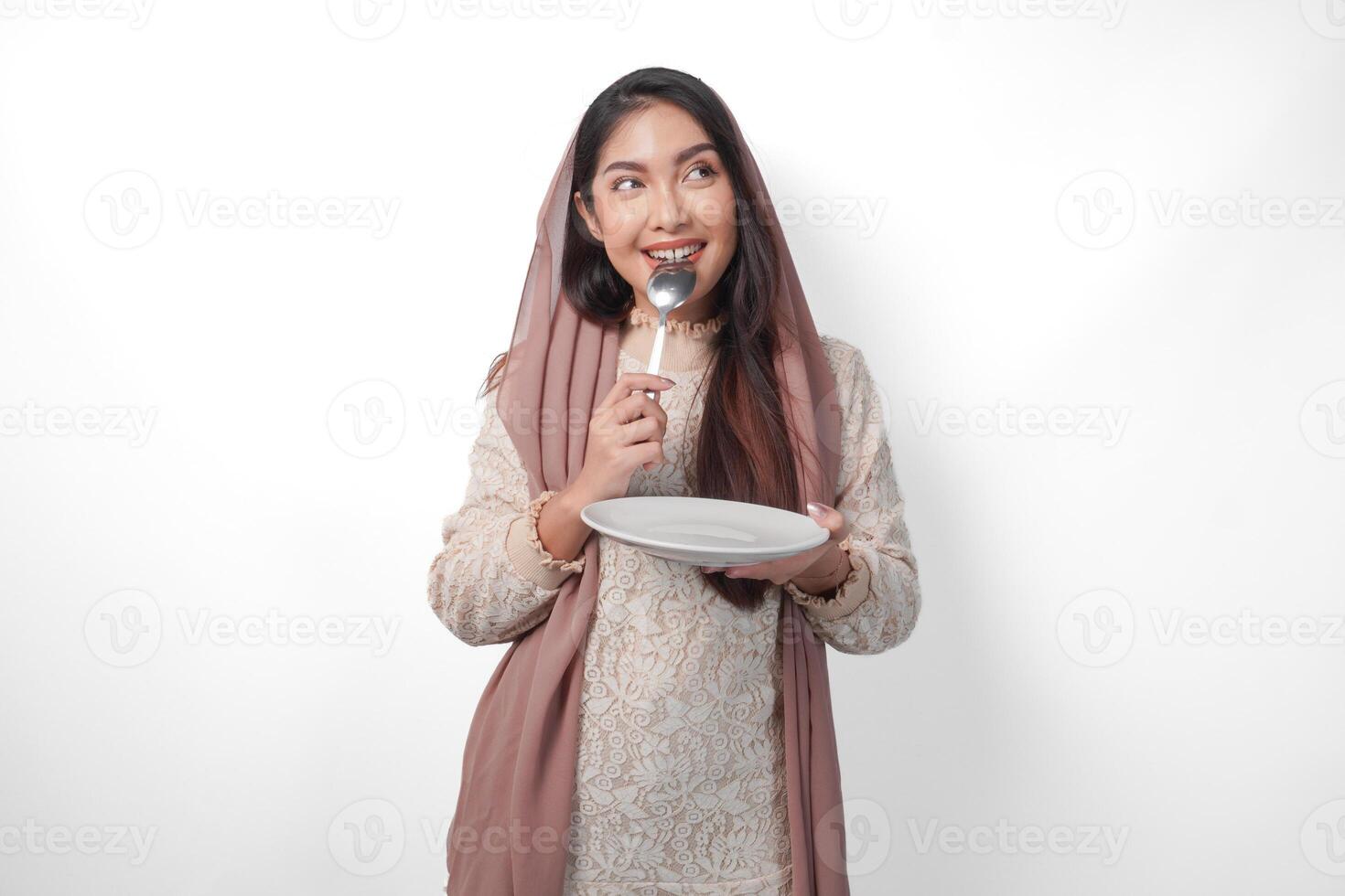 hongerig Aziatisch moslim vrouw Holding bord denken wat naar eten met kopiëren ruimte over- de bord terwijl bijten lepel aan het wachten voor iftar, geïsoleerd wit achtergrond foto