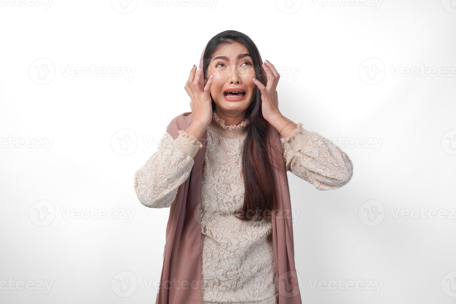 een Aziatisch moslim vrouw verhogen beide handen naar houden haar hoofd gevoel benadrukt en beduusd, huilen en schreeuwen wanhopig, geïsoleerd wit achtergrond foto