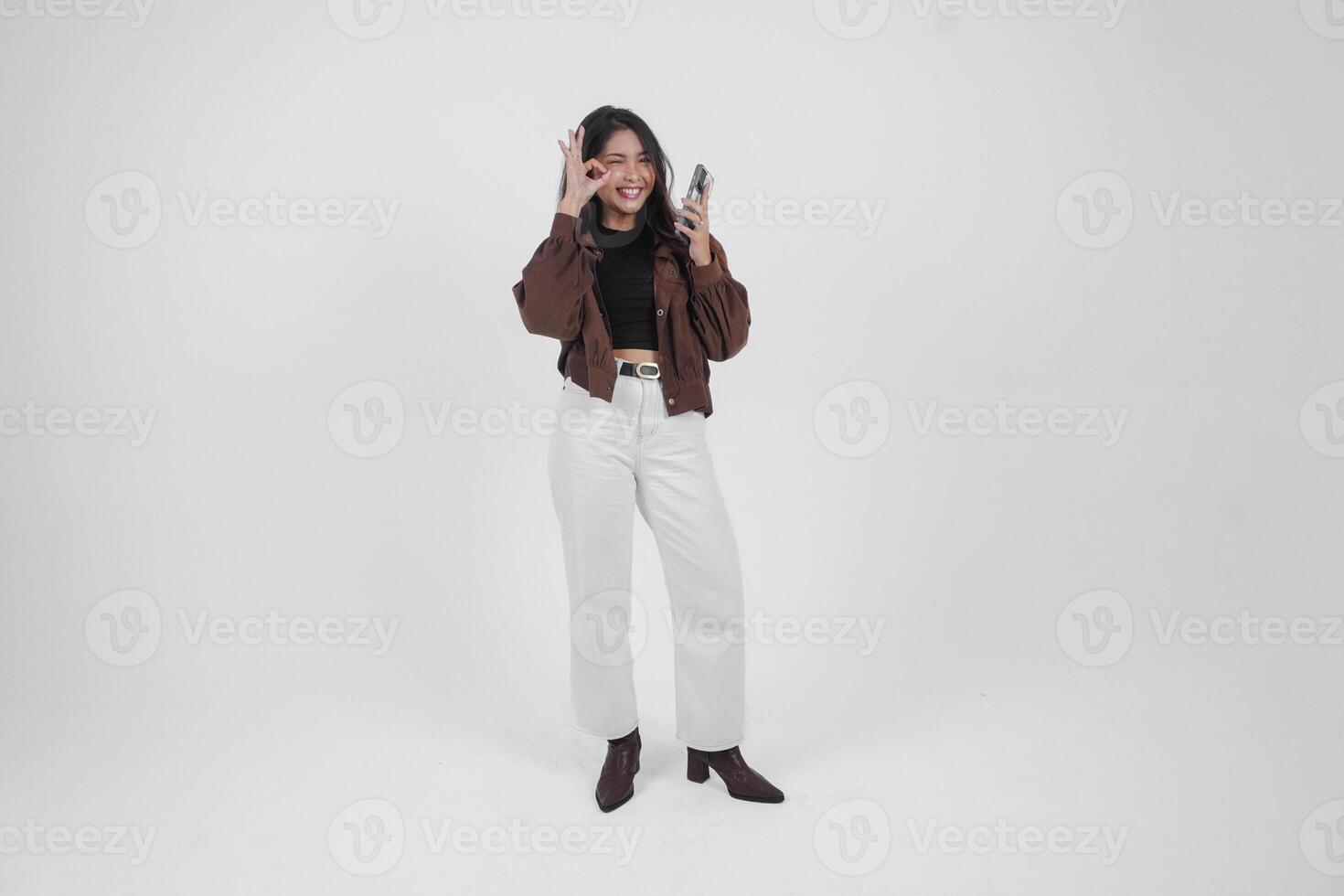 opgewonden jong Aziatisch vrouw tonen Oke teken met Rechtsaf hand- en Holding telefoon gebruik makend van links hand, vervelend gewoontjes kleren Aan geïsoleerd wit achtergrond foto