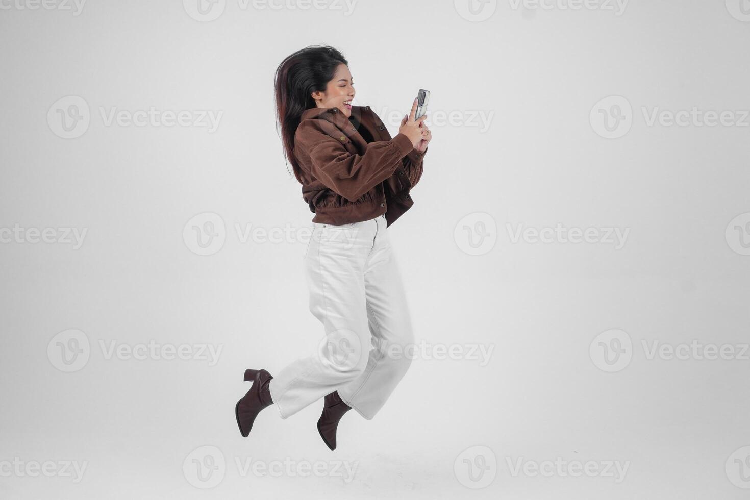 vrolijk Aziatisch vrouw jumping en glimlachen in lucht terwijl Holding en op zoek Bij telefoon, gevoel opgewonden met een breed lach Aan geïsoleerd wit achtergrond foto