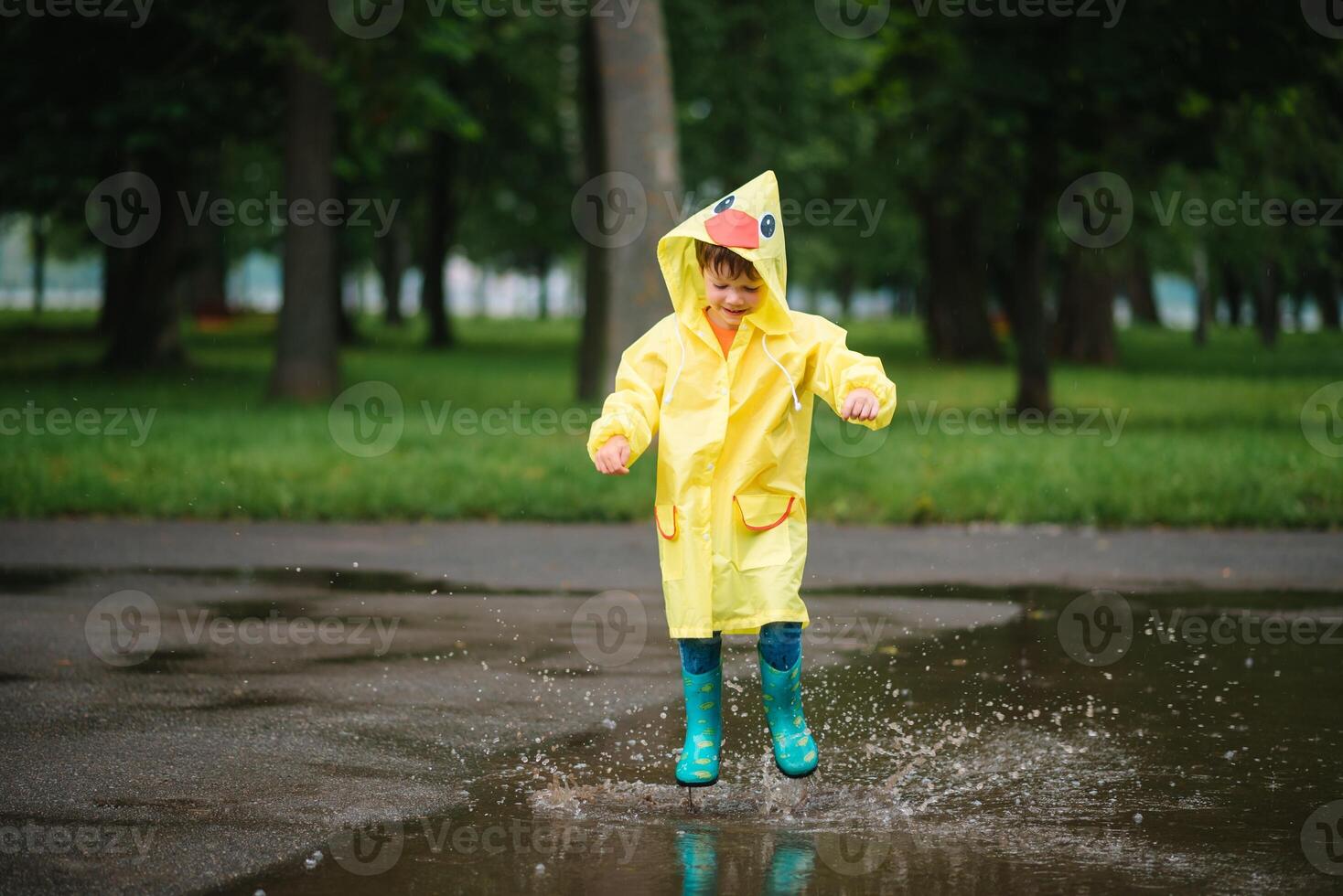 weinig jongen spelen in regenachtig zomer park. kind met paraplu, waterbestendig jas en laarzen jumping in plas en modder in de regen. kind wandelen in zomer regen buitenshuis pret door ieder het weer. gelukkig jeugd. foto