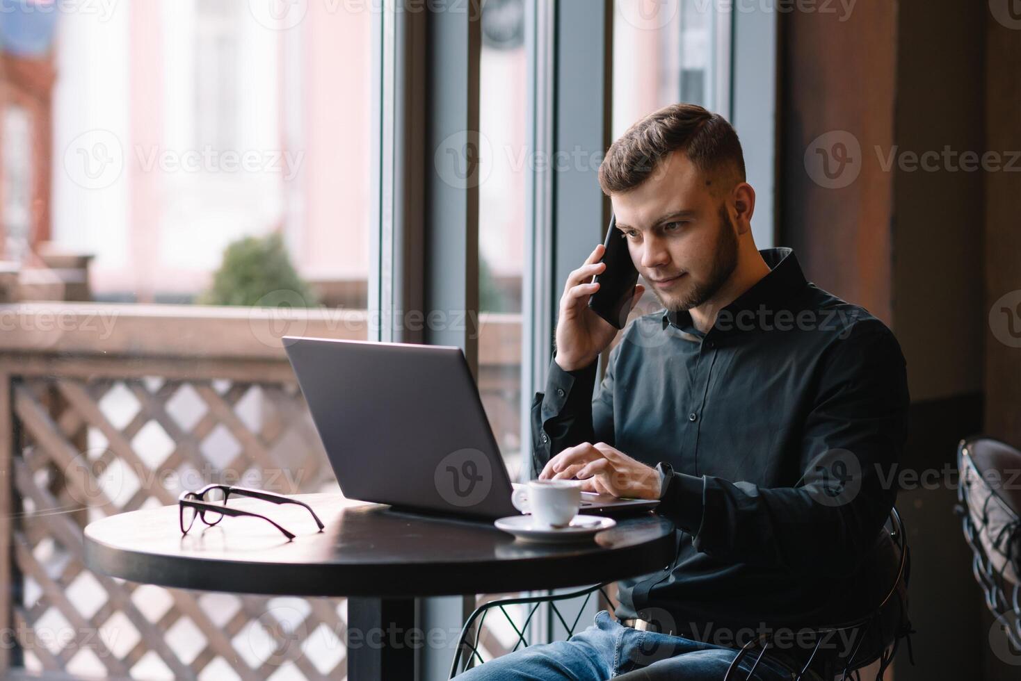 jong zakenman pratend Aan mobiel telefoon terwijl werken Aan laptop in cafe. foto